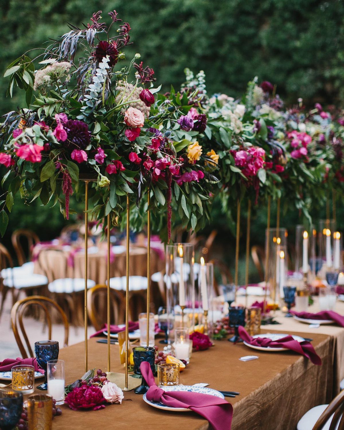 Floral centerpieces on tall gold stands decorate a long wooden wedding reception table.