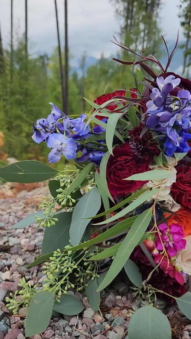 Deep red roses and purple irises with green eucalyptus leaves resting on gravel outdoors.
