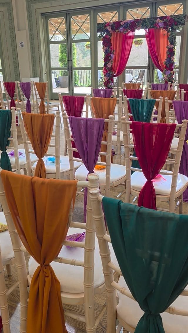 White chairs decorated with colorful jewel-toned fabric sashes in a wedding ceremony setting.
