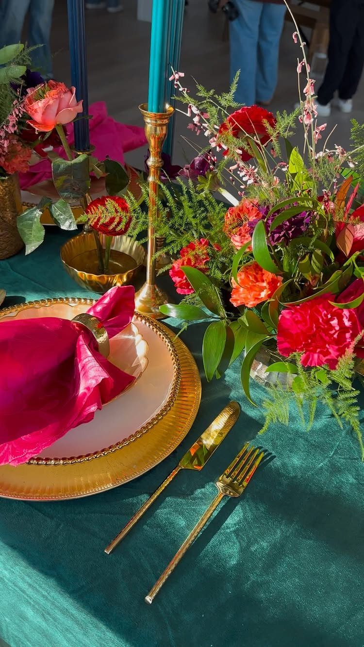 A formal table setting with a gold charger, magenta napkin, and colorful floral centerpiece on a teal tablecloth.