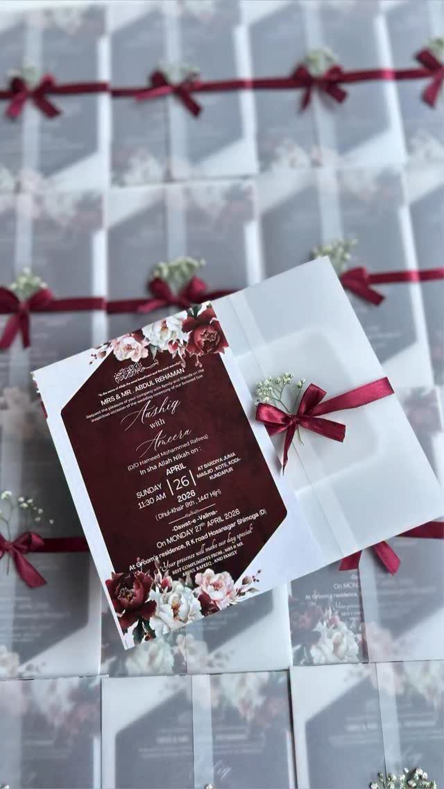 Wedding invitation card with burgundy floral patterns resting on white envelopes tied with red ribbons.