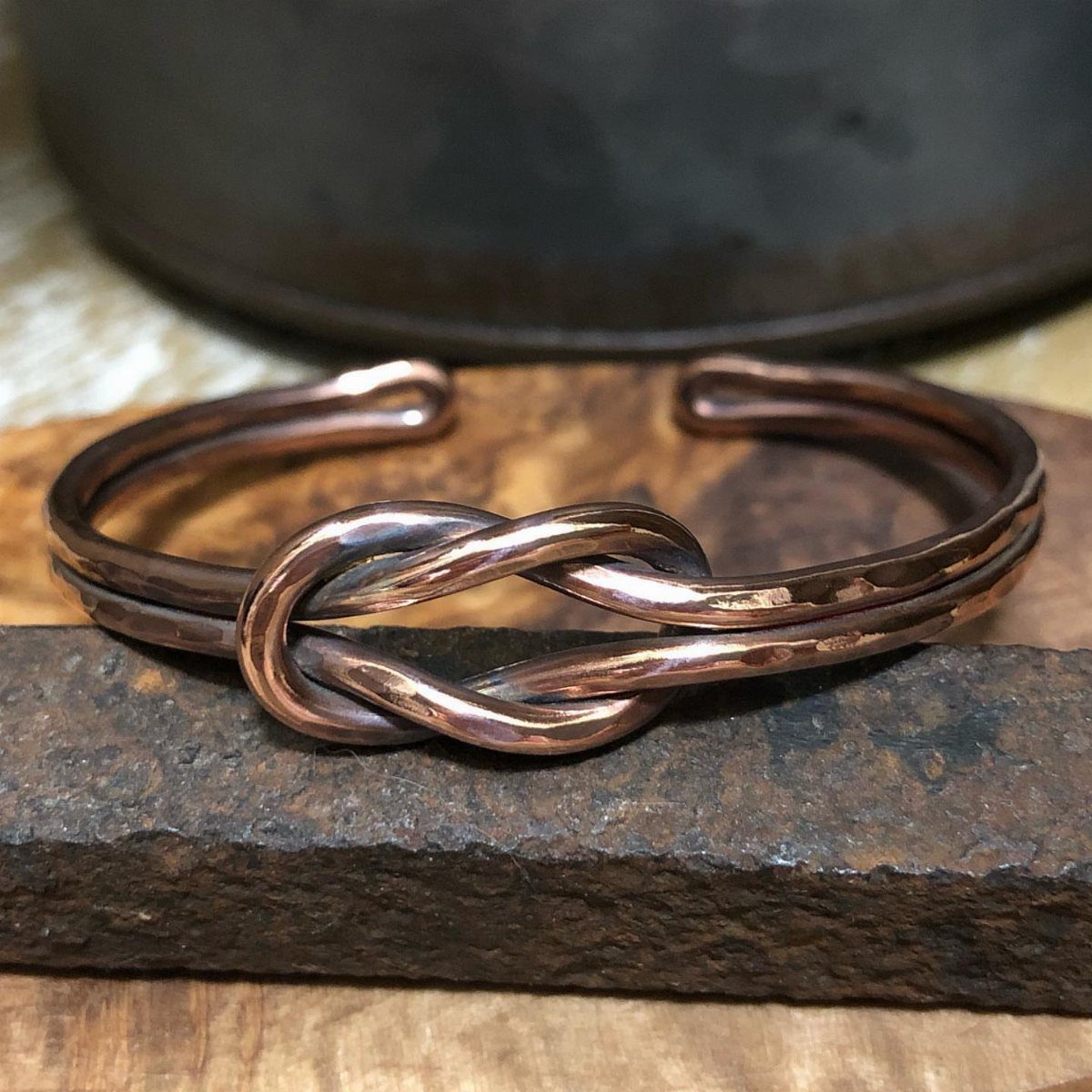 A hammered copper cuff bracelet with a prominent knot design resting on a dark wooden surface.