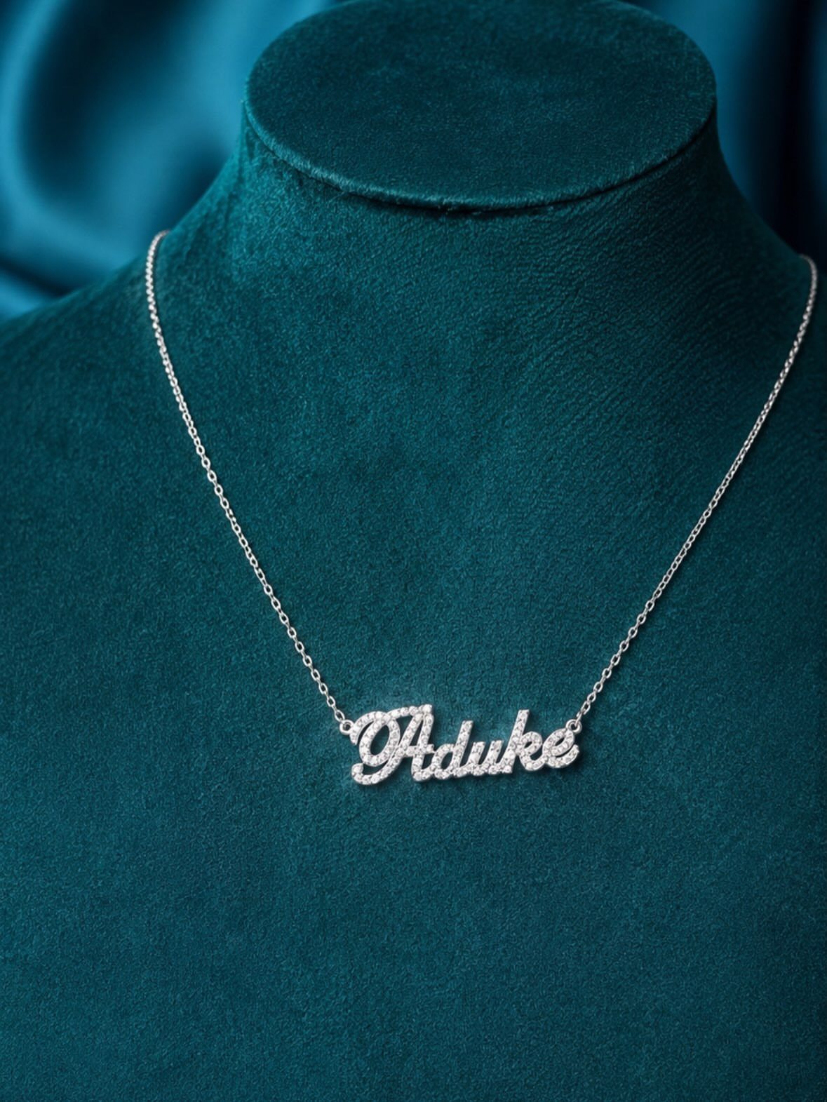 A silver name necklace with sparkling stones displayed on a teal velvet jewelry bust.
