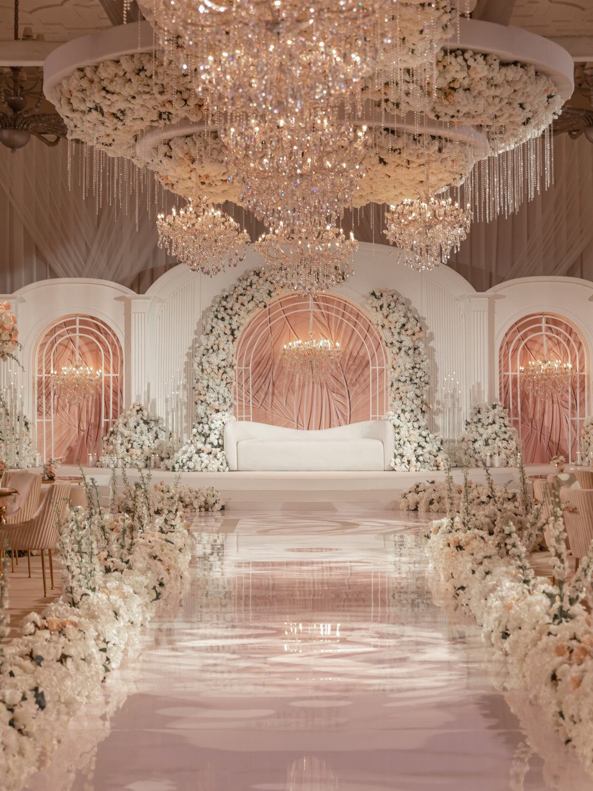 A glossy pink wedding aisle lined with white and light pink flowers under large crystal chandeliers.