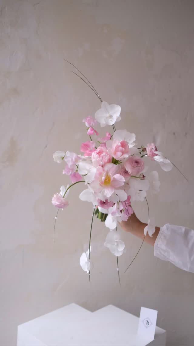 A hand holding a delicate bouquet of light pink and white flowers against a neutral background.