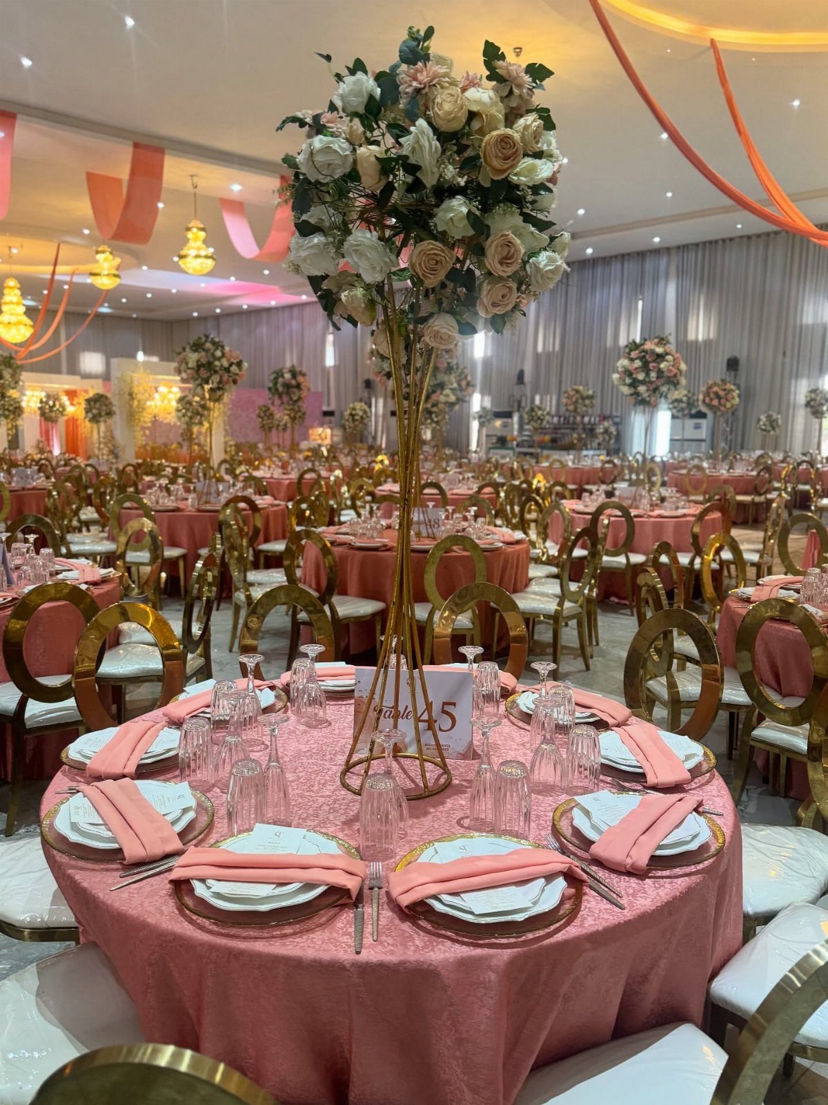 A round wedding table set with pink linens, gold chargers, and a tall floral centerpiece.