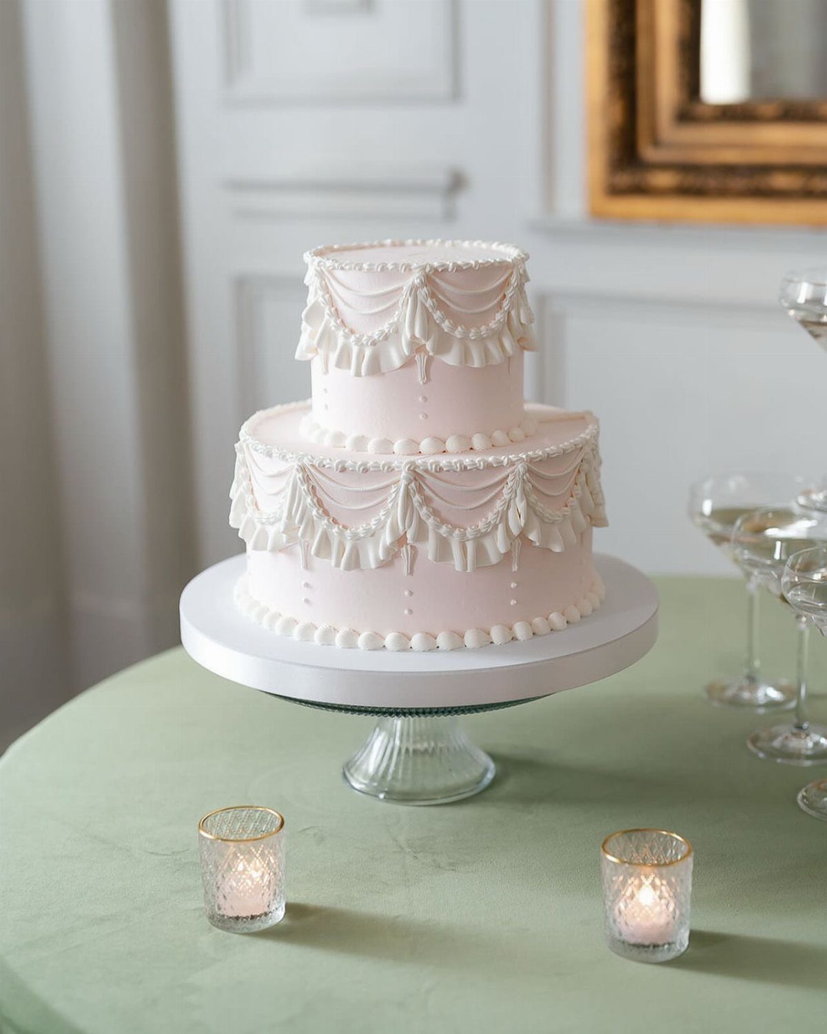A two-tier light pink wedding cake with white frosting swags on a sage green tablecloth.