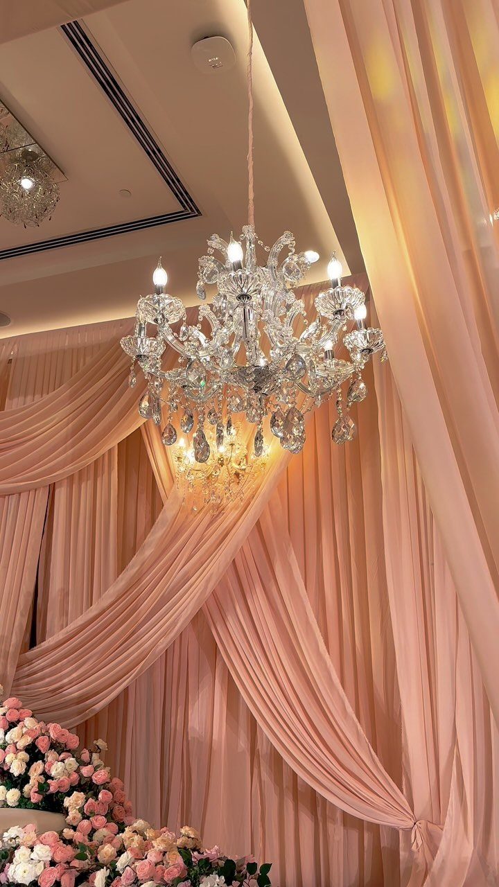 Crystal chandelier hanging against draped light pink fabric and pink floral arrangements.