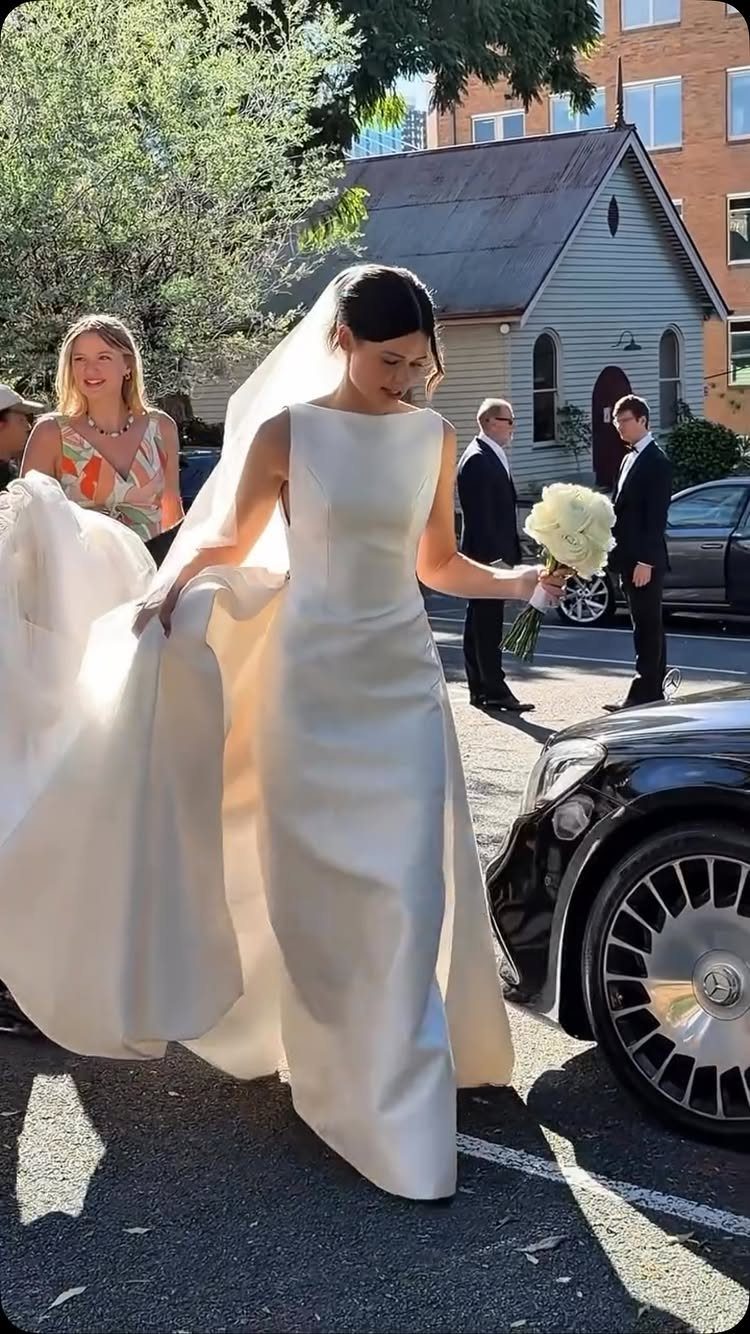 A bride in a minimal white satin wedding dress walks while holding white roses.