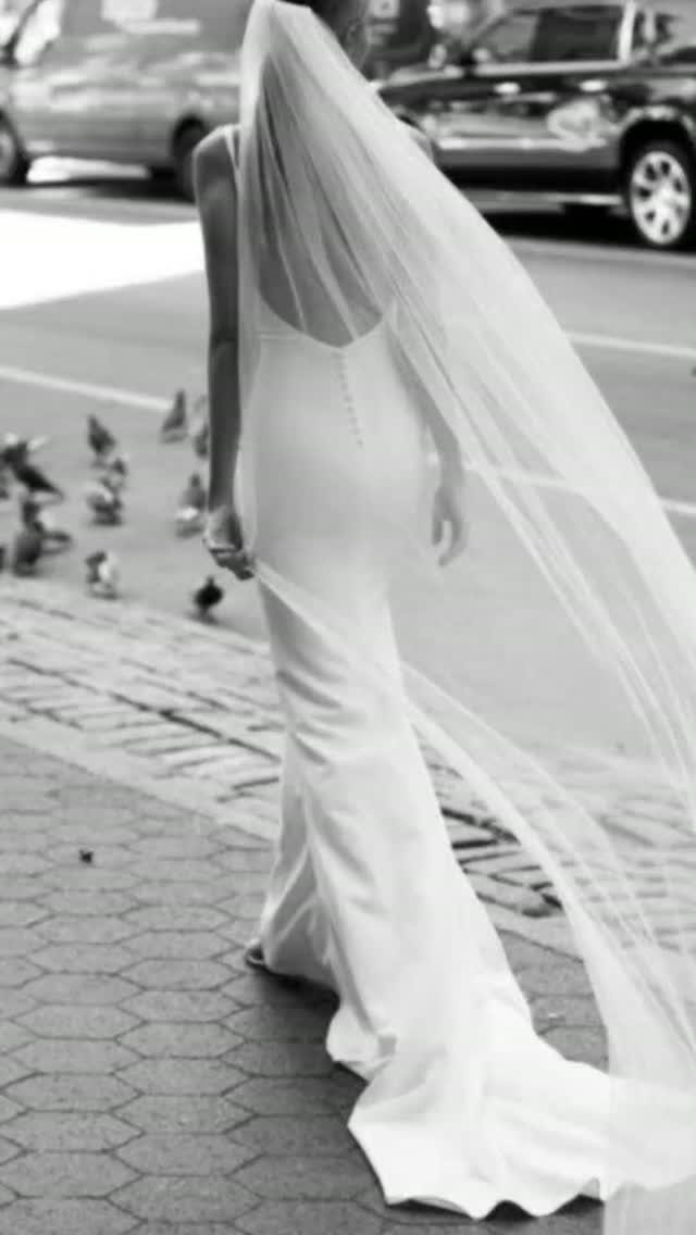 A woman wearing a white slip wedding dress and a long sheer veil walking on a city street.