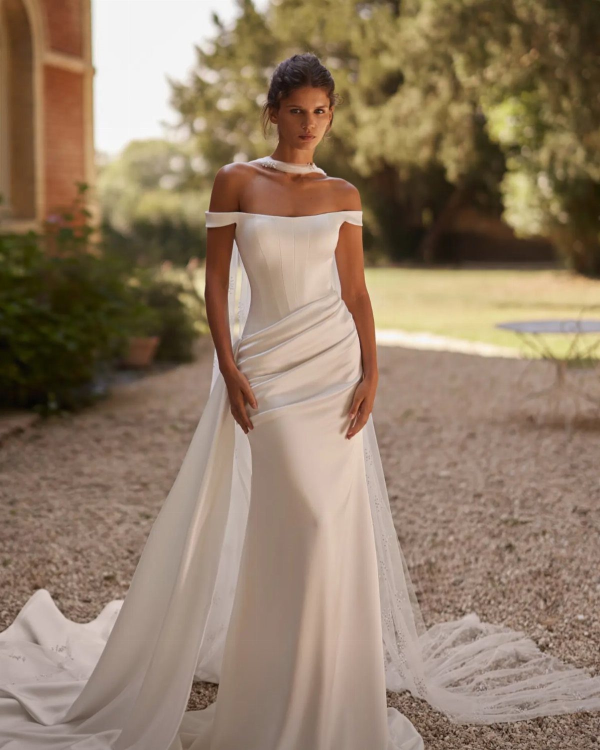 A woman in a minimal off-the-shoulder white wedding dress standing on a gravel path.