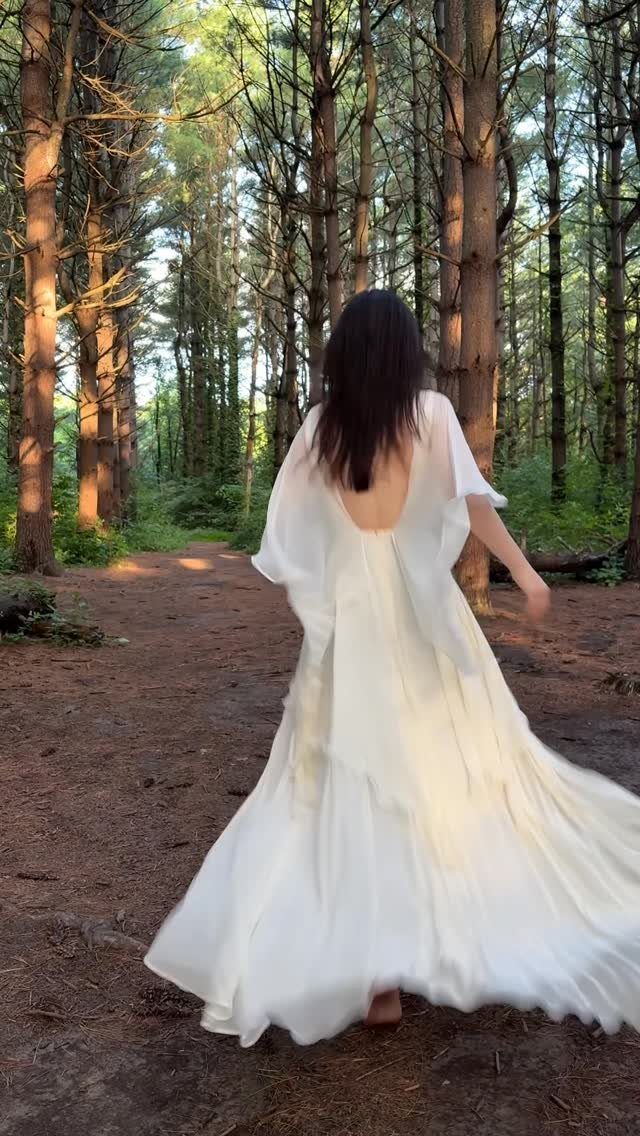 A woman in a flowing white minimal wedding dress walking through a sunlit forest.