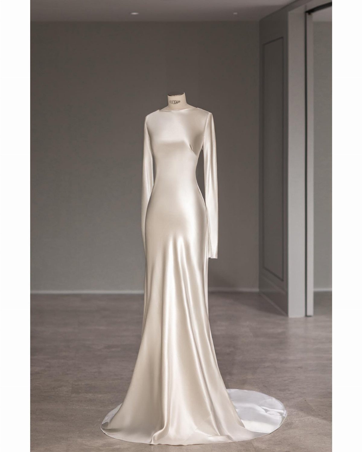 A long-sleeved ivory silk wedding dress displayed on a mannequin in a minimalist room.