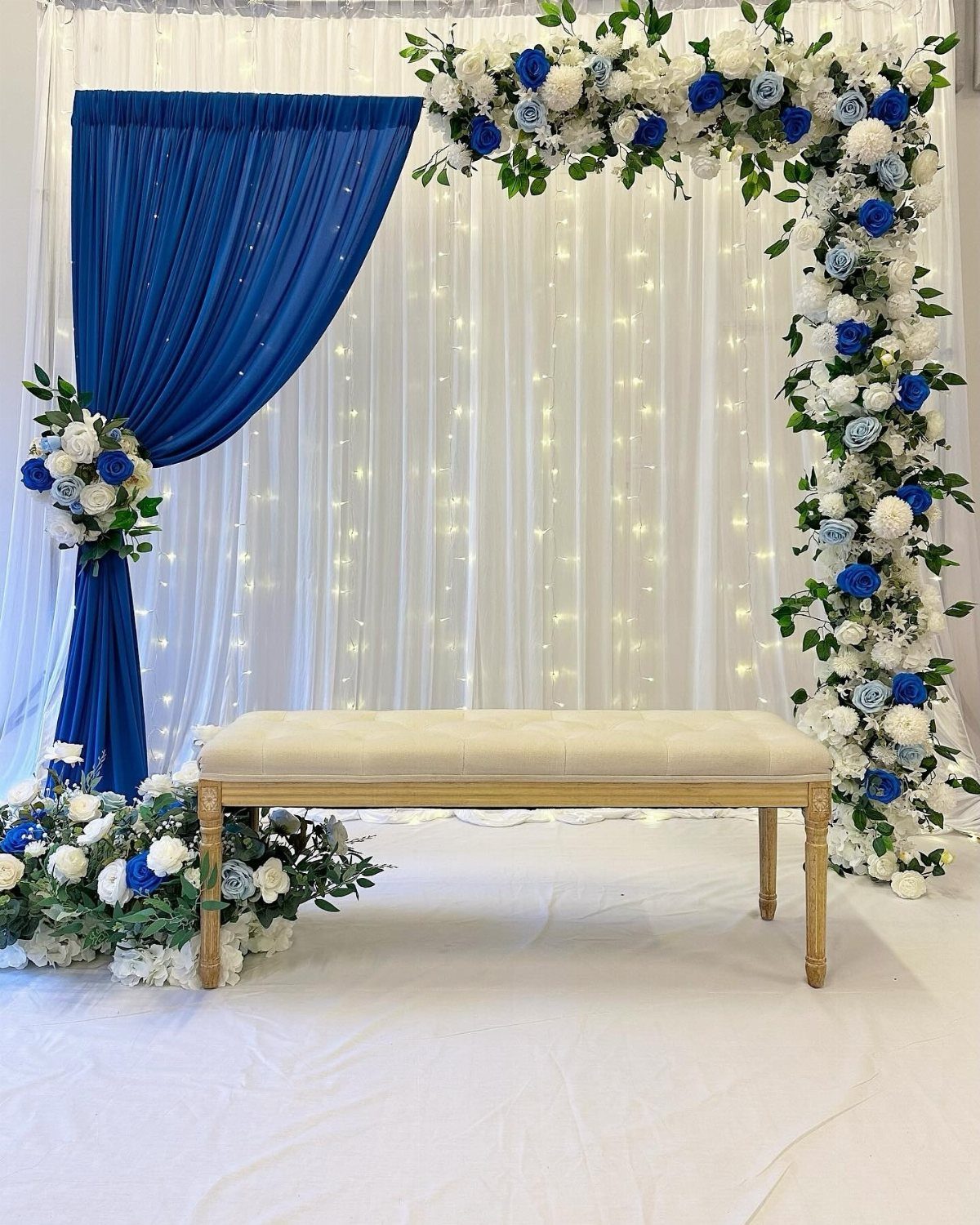 Wedding backdrop with navy blue drapery, white fairy light curtains, and blue and white floral arrangements.