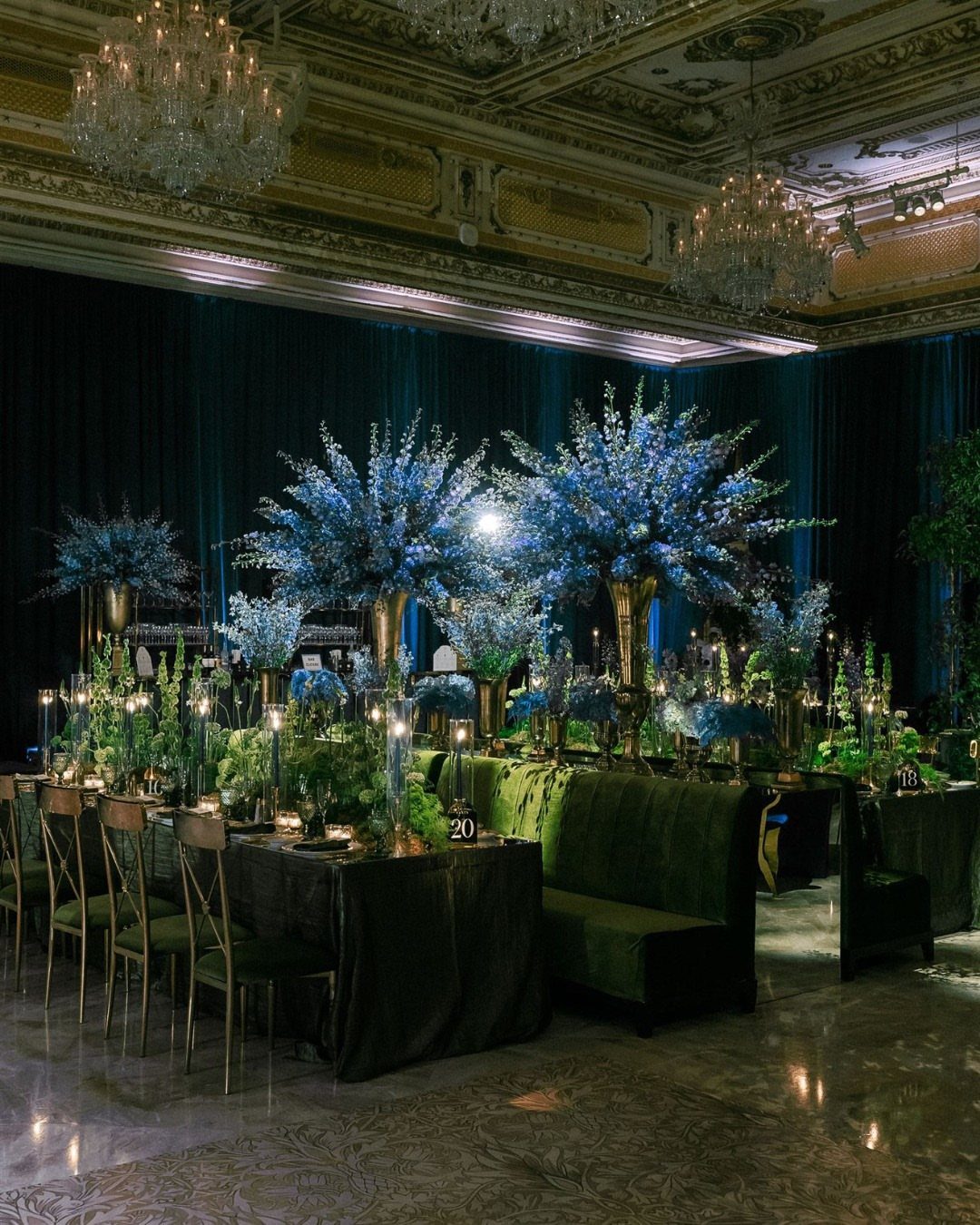 Blue floral centerpieces on tables with green velvet seating in a grand ballroom.