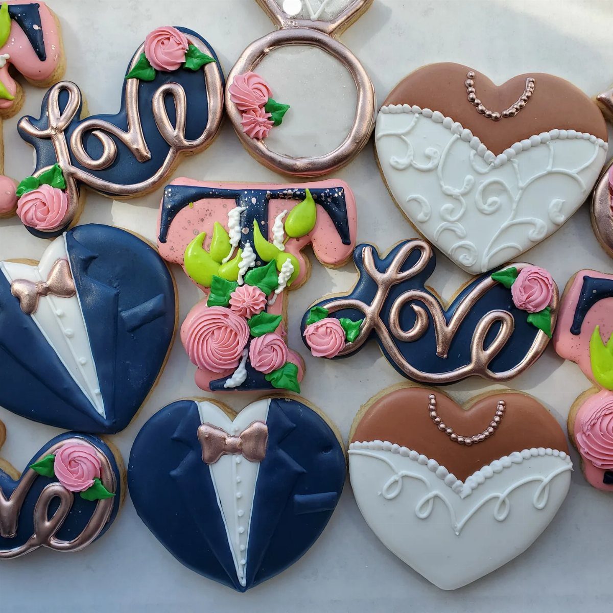 Decorated sugar cookies with navy blue, pink, and rose gold icing in various wedding shapes.