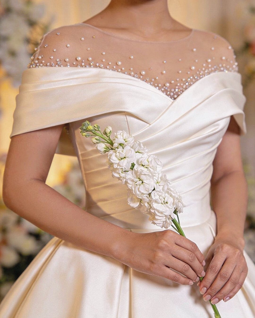 A person in an off-white satin wedding dress with pearl-embellished sheer neckline holding white flowers.