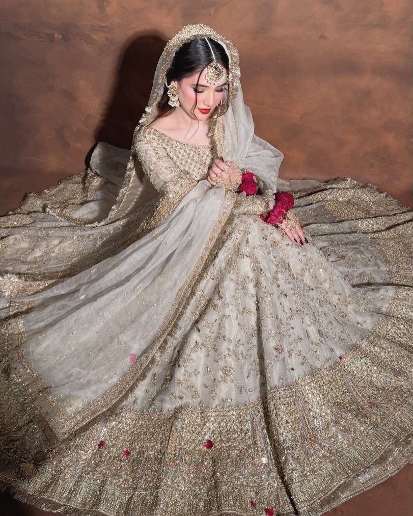 Woman in an ornate off-white and gold embroidered wedding lehenga sitting against a brown backdrop.