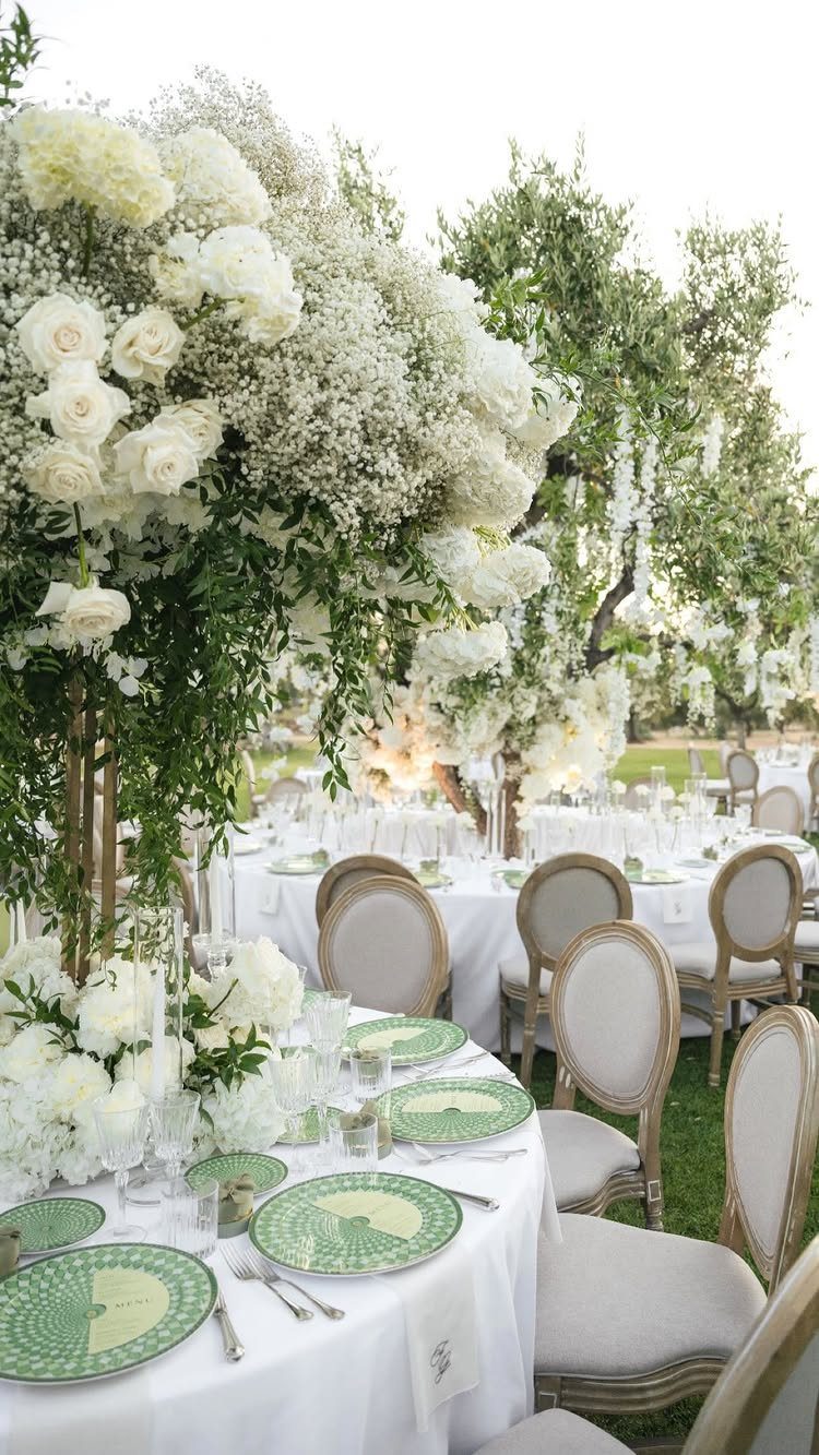 Wedding reception tables set with green patterned plates and white floral arrangements under trees.