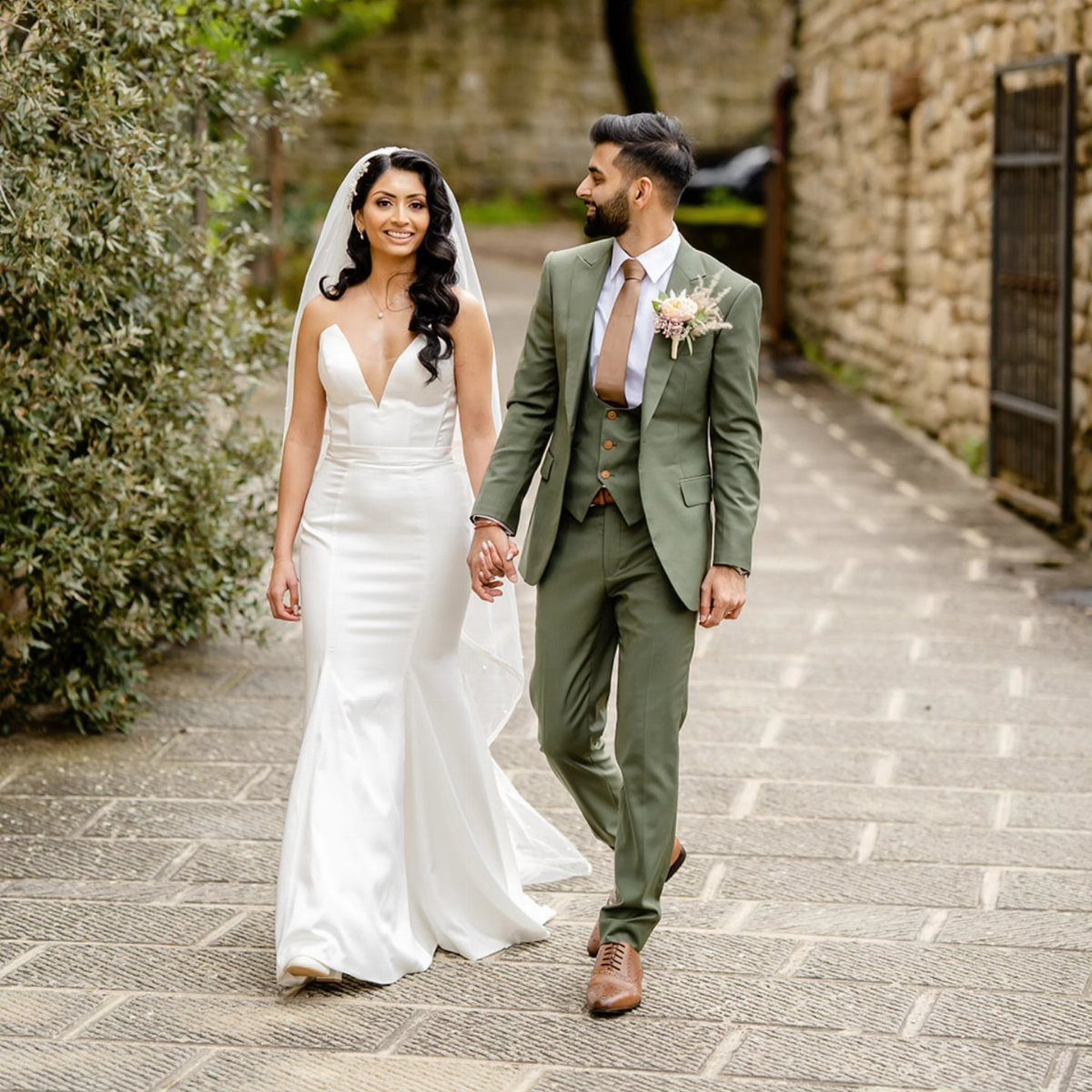 A bride and groom in an olive green suit walking together on a stone path.