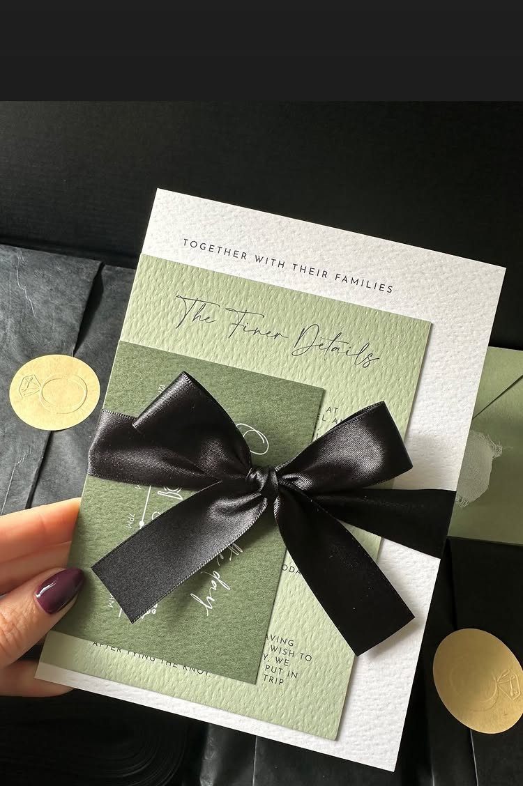 Hand holding olive green wedding invitations tied with a large black satin bow and gold wax seals.