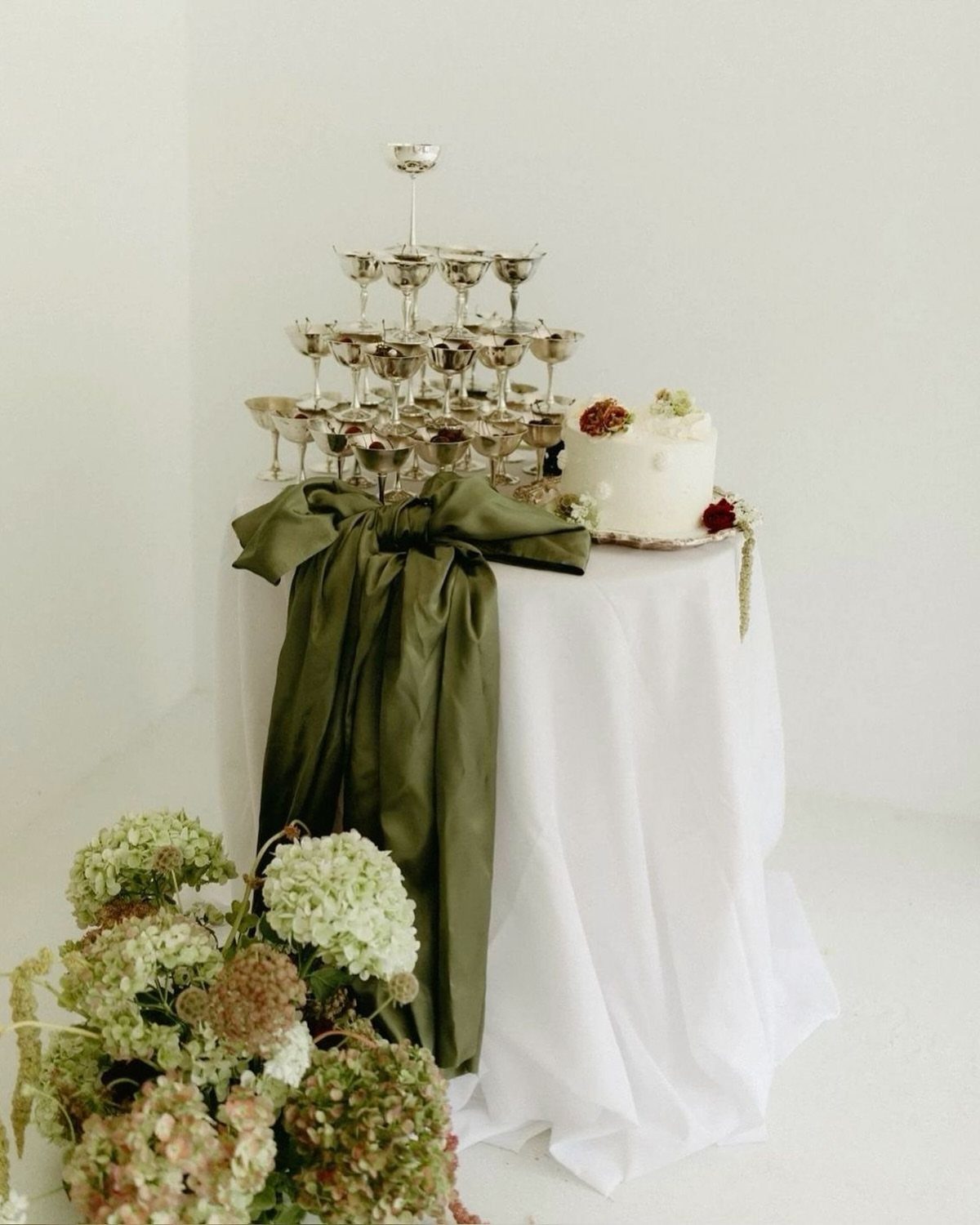 A white table featuring an olive green satin bow, a small cake, and a silver champagne tower.