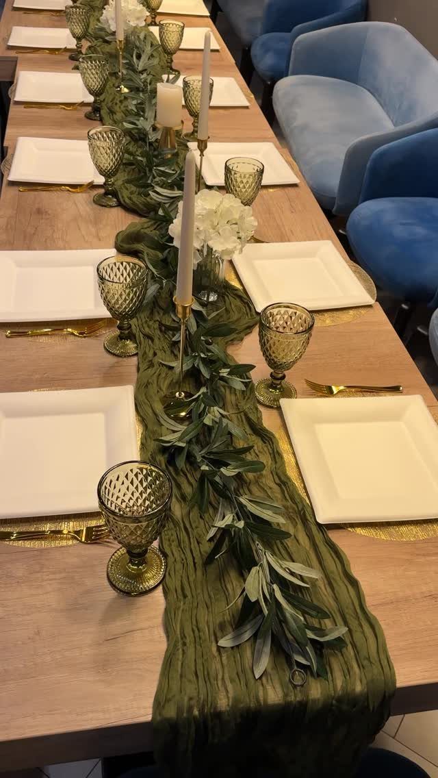 A long wooden table decorated with olive leaves, green glassware, white plates, and gold cutlery.