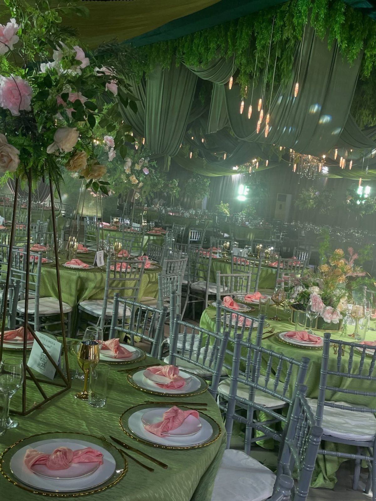 Wedding reception tables with olive green linens, pink napkins, and silver chairs under draped ceilings.