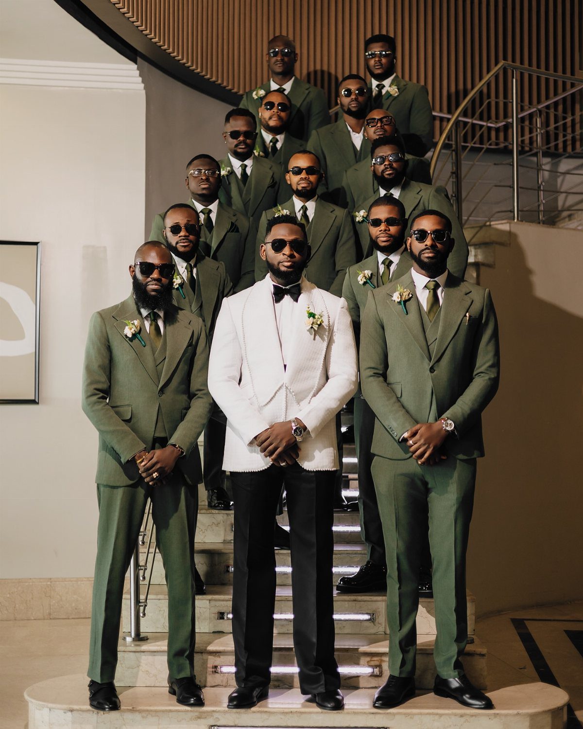 Men in olive green suits and formal attire posing together on a staircase.