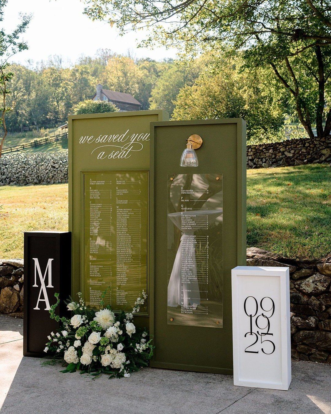 Two olive green wedding seating chart signs standing outdoors near white flowers.