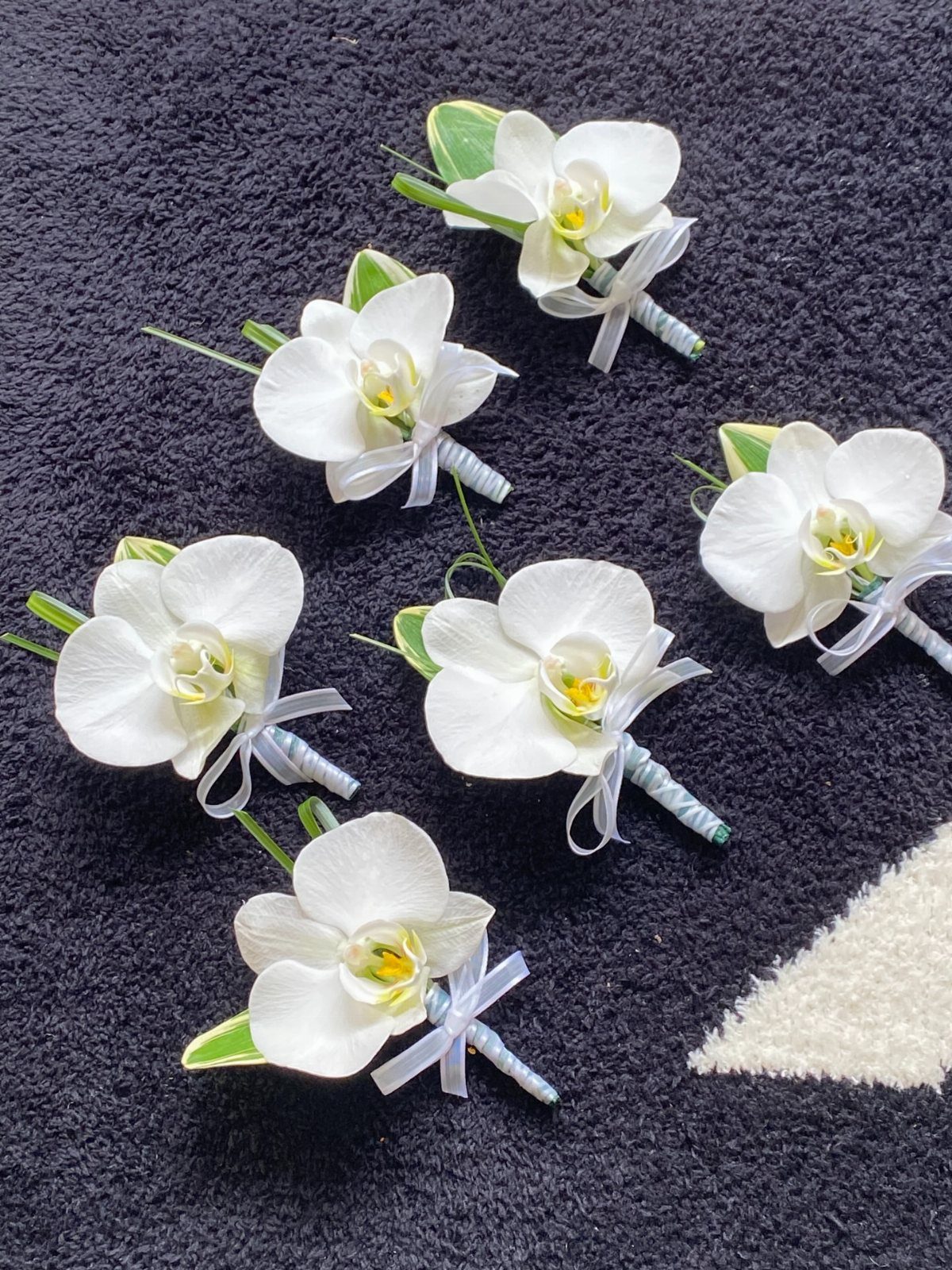 7 Orchids Bouquet Wedding Ideas for Every Style of Ceremony 4 Six white orchid boutonnieres with green leaves arranged on a dark textured fabric background.