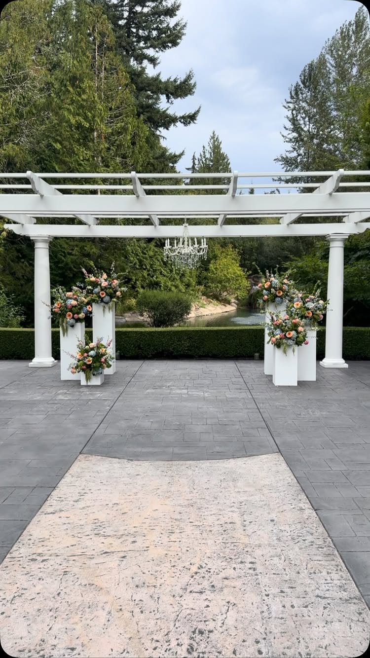 Outdoor Wedding Altar: 5 Ideas That Cost Less Than They Look 4 White pillars with peach and blue floral arrangements under a white pergola on a stone patio.
