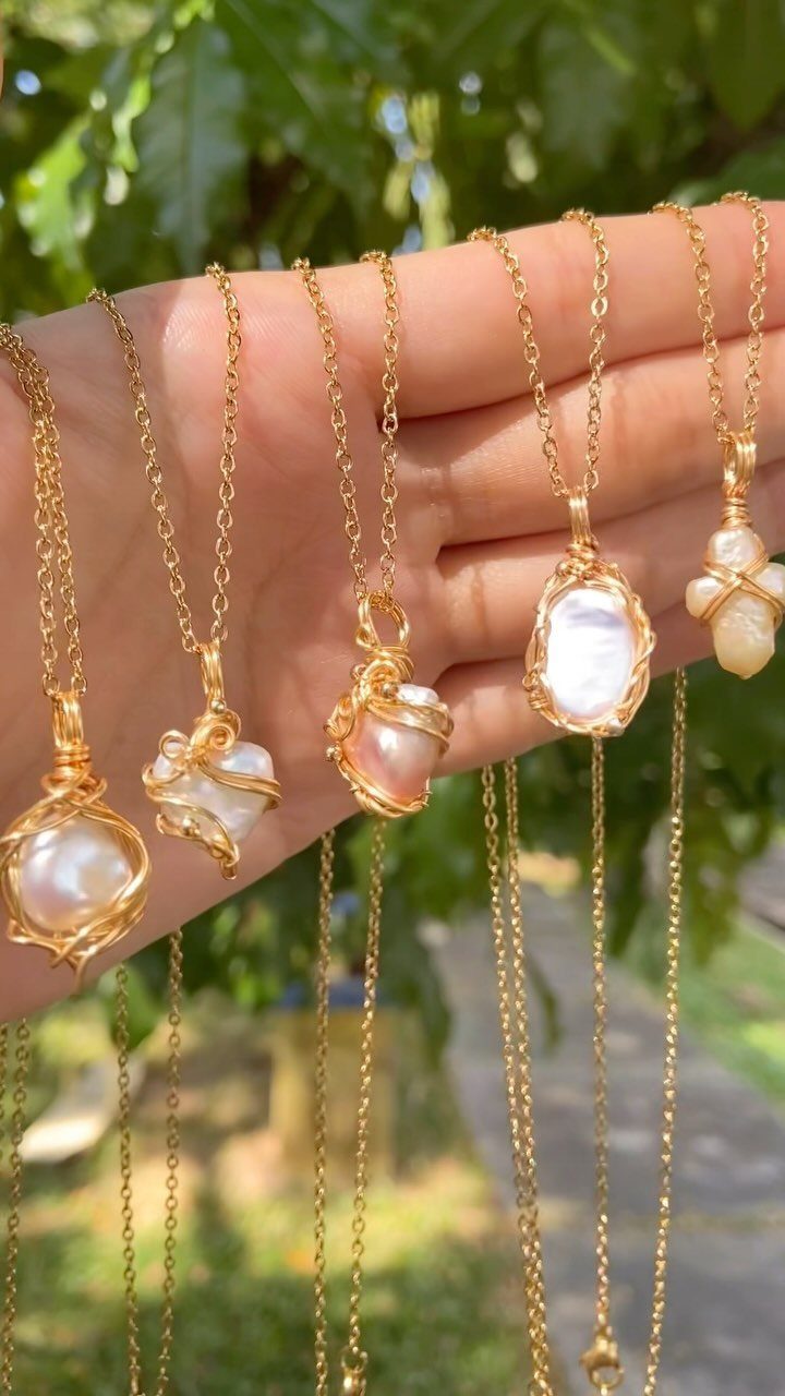 Hand holding five gold necklaces with irregular pearl pendants against a green background.