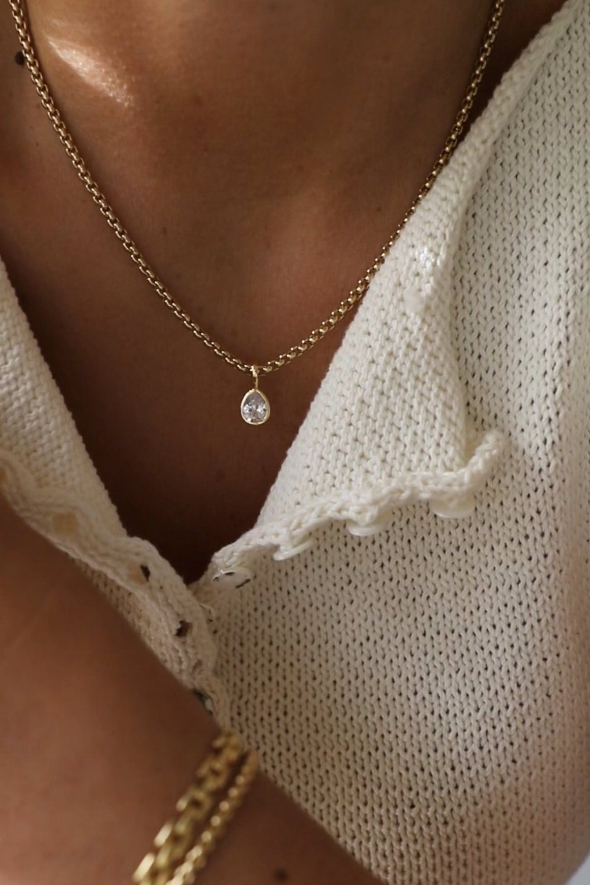 A delicate gold chain necklace with a small clear pendant worn against a white knit shirt.