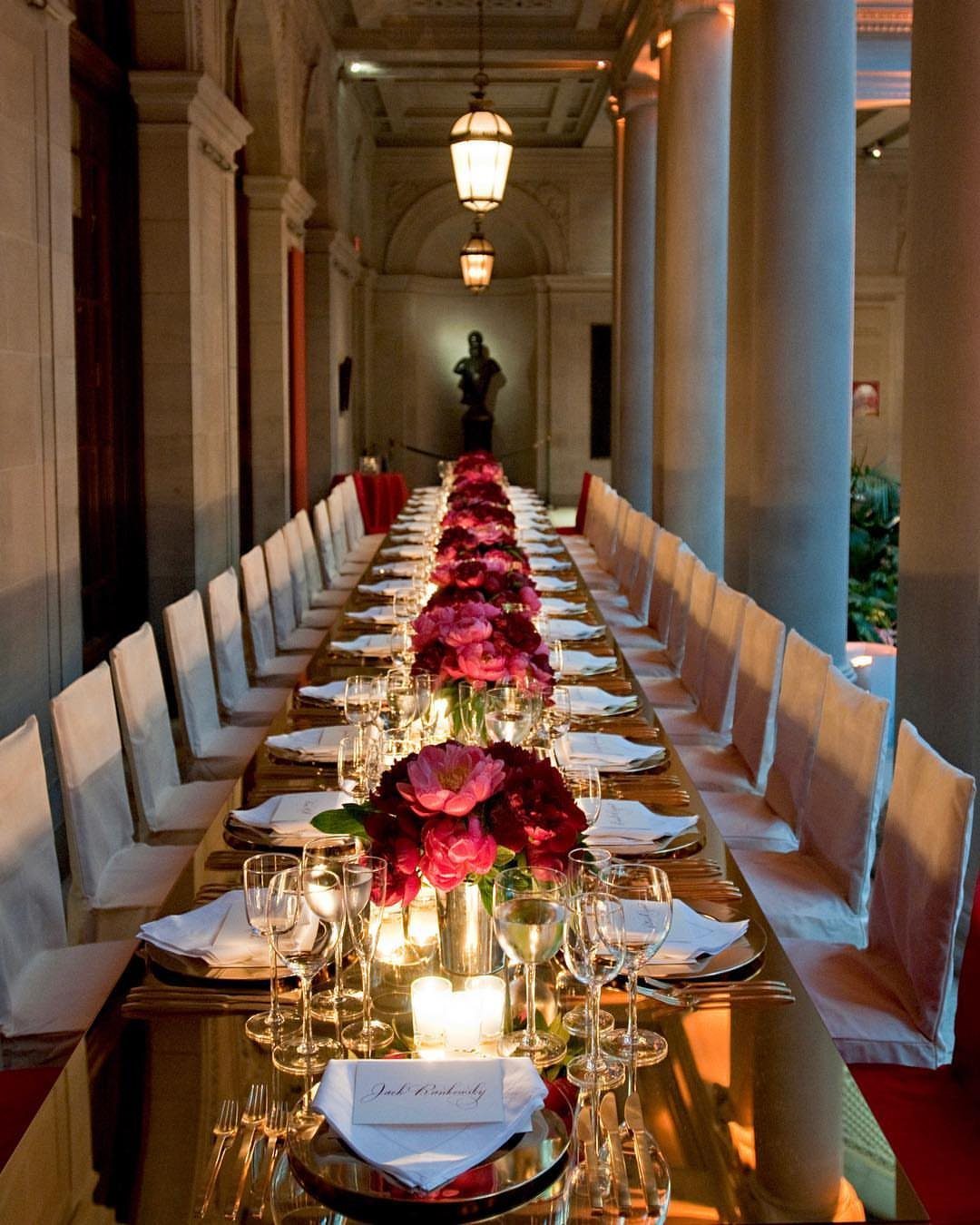 A long banquet table with pink flowers set inside a grand hall with stone columns.