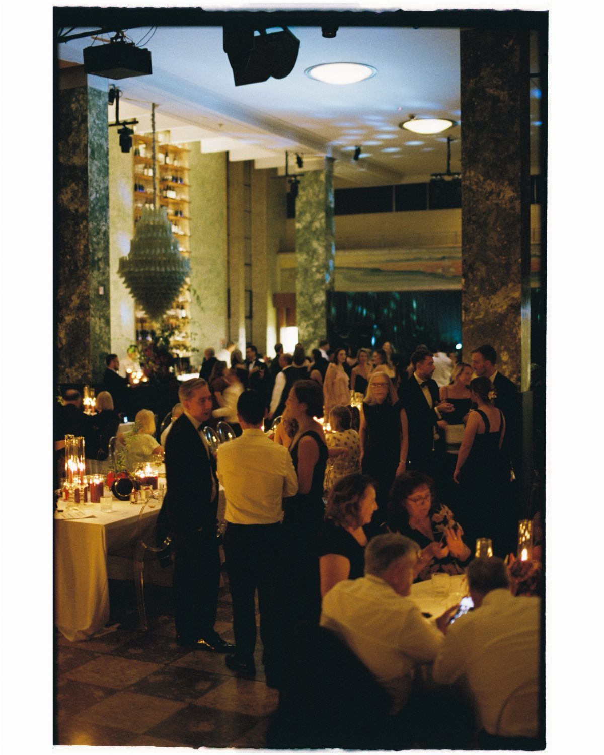 People gathered for a formal reception in a large hall with marble columns and a chandelier.