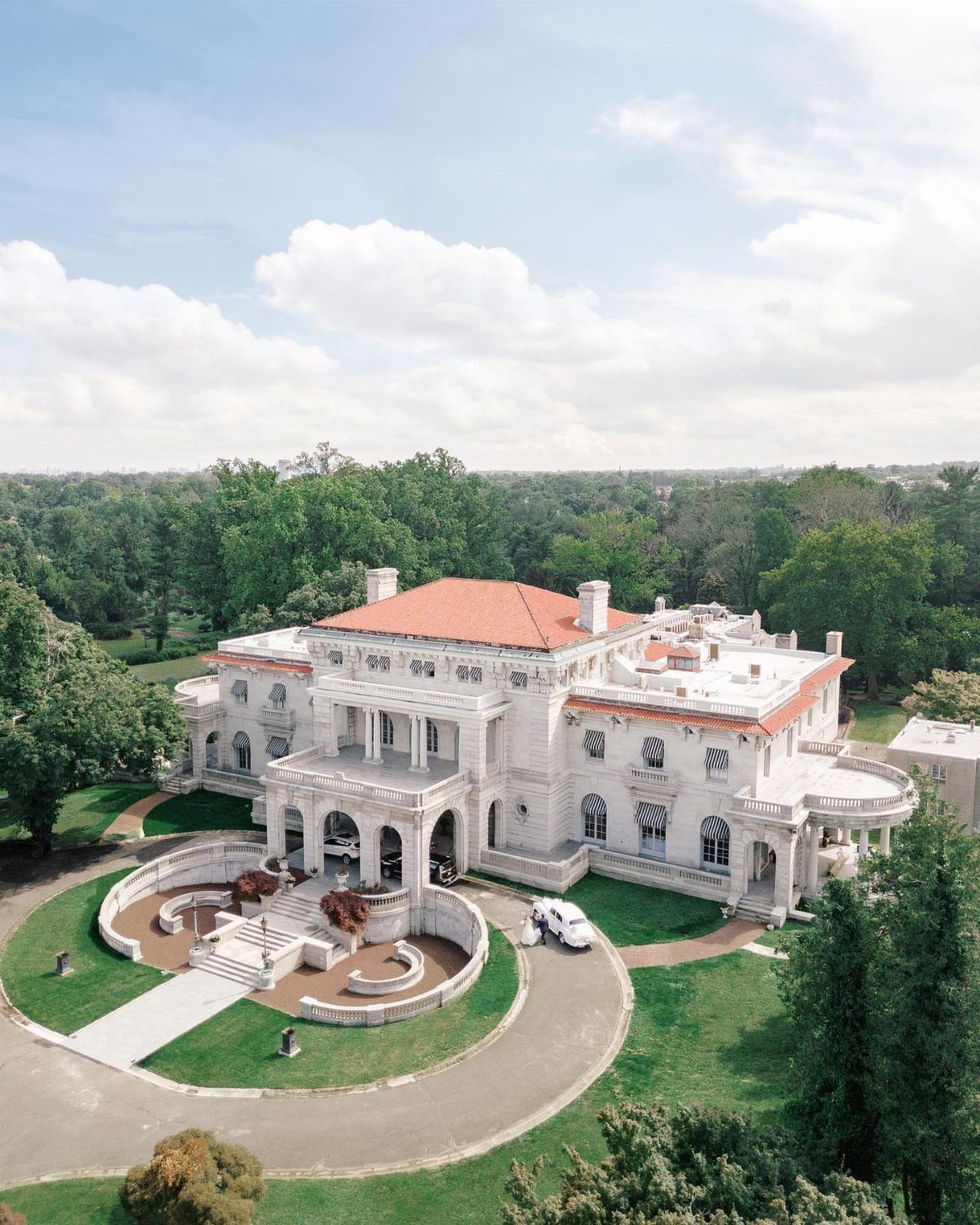 A large white historic mansion with a red tile roof surrounded by green trees and lawns.
