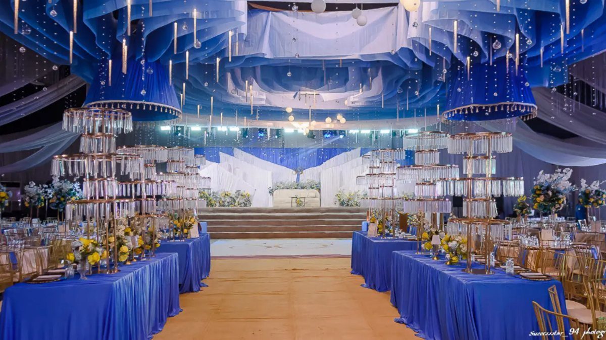 A wedding reception hall featuring blue draped ceilings, blue tablecloths, and tall glass centerpieces.