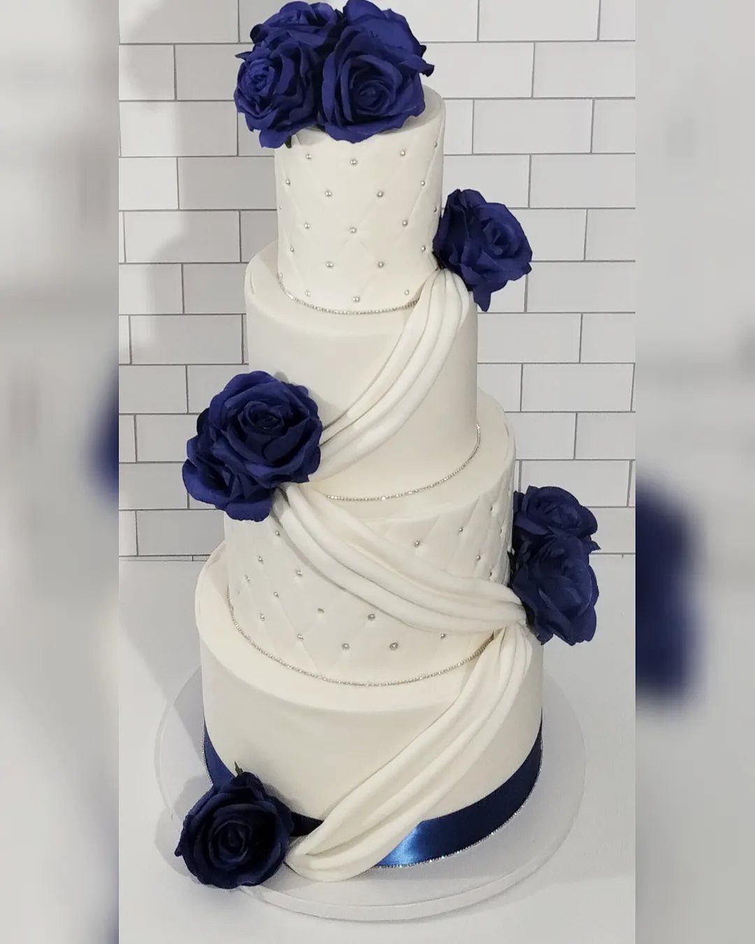 A four-tier white wedding cake decorated with royal blue roses and silver pearls.