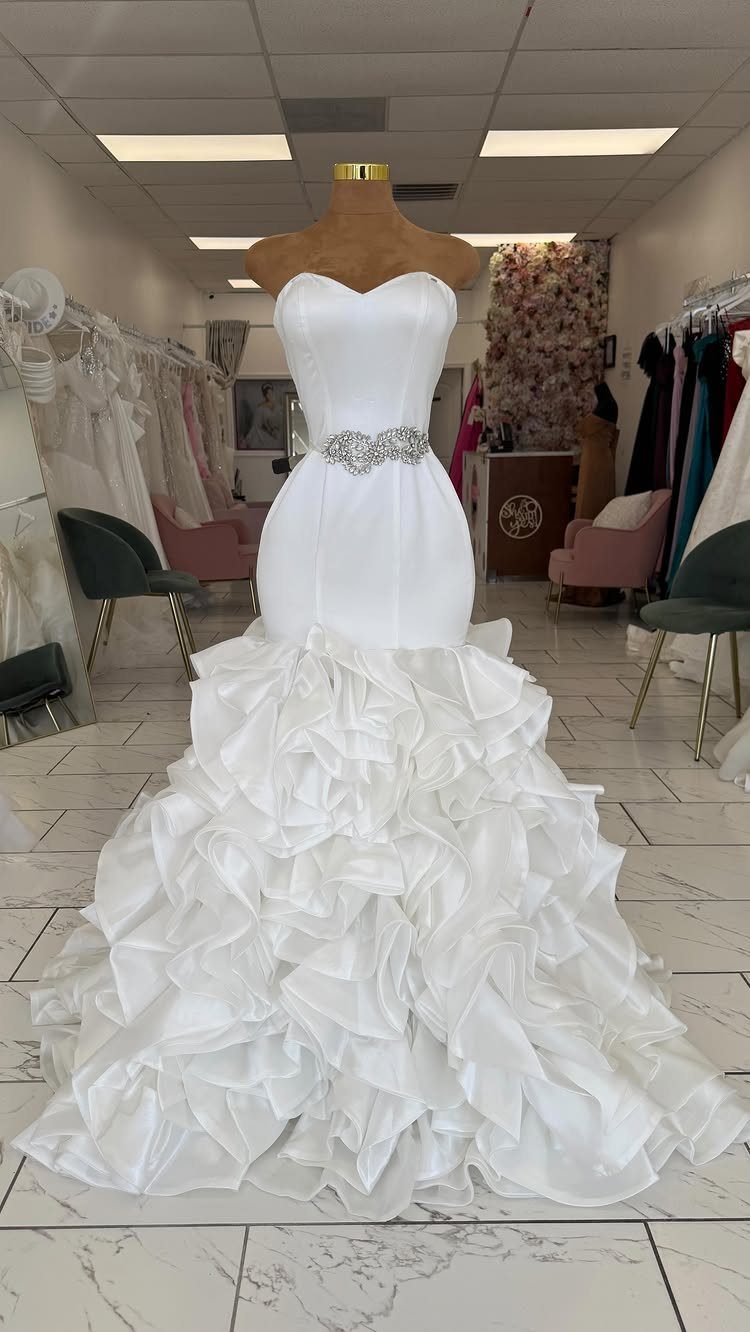 A white mermaid wedding dress with a sparkling belt and voluminous cascading ruffles on a mannequin.