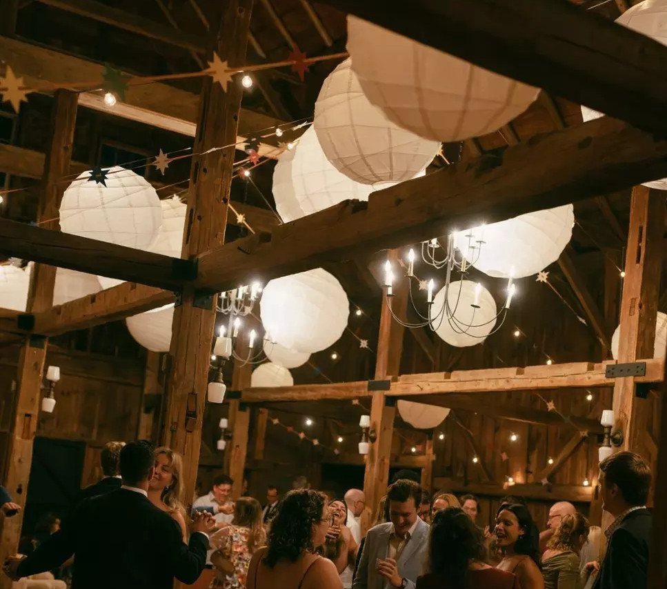 White paper lanterns hanging from wooden barn beams above a crowded wedding reception.