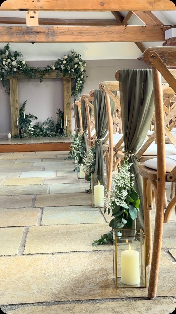 Wooden chairs with green fabric and glass candle lanterns lining a stone wedding aisle.