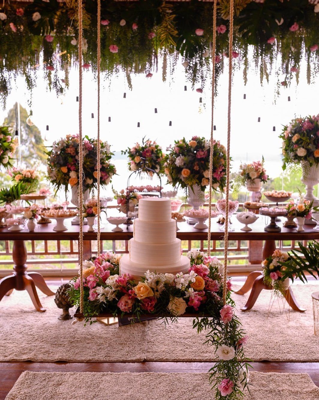 A white wedding cake on a flower-decorated swing suspended by ropes outdoors.