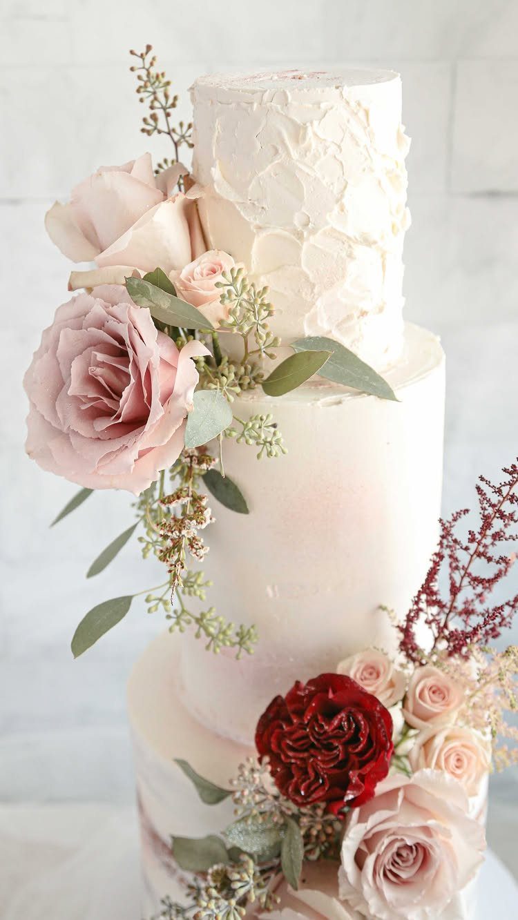 A three-tiered white wedding cake decorated with various pink and red roses and greenery.