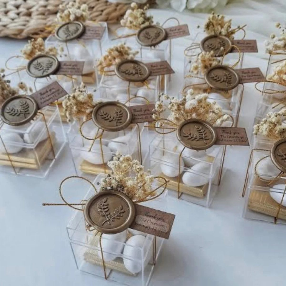 Small clear favor boxes with white candies, dried flowers, and wax seals on a white surface.
