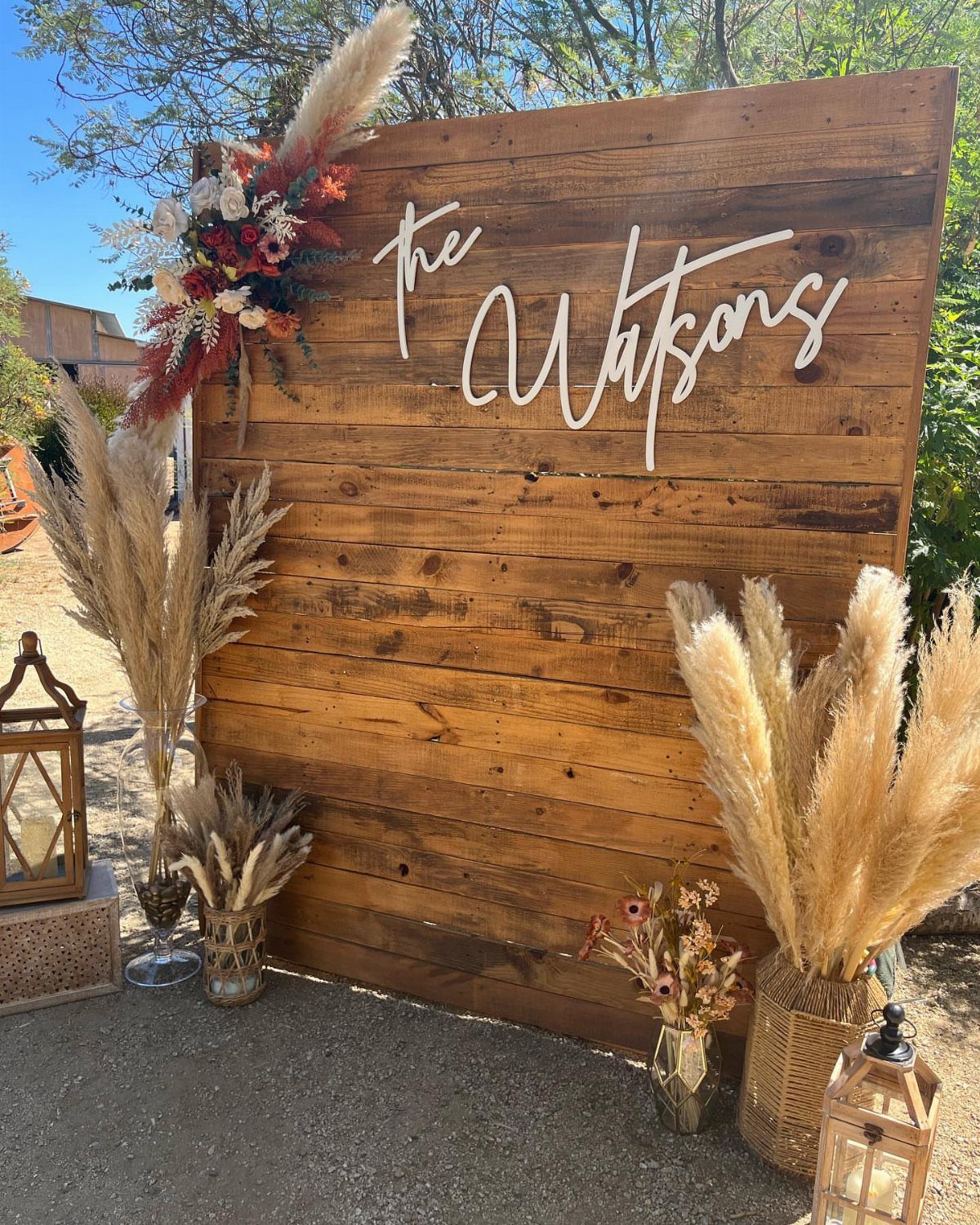 Wooden plank backdrop with white cursive text decorated with pampas grass and flowers.