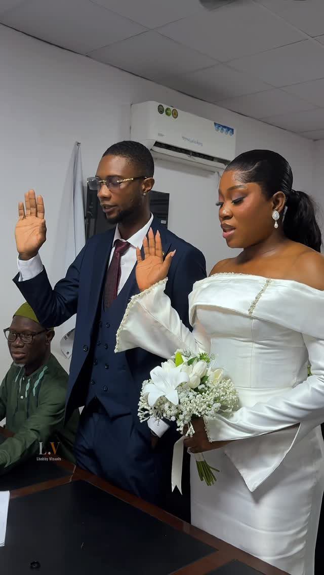 Simple Court Wedding Dress: 7 Styles That Work at City Hall 3 A bride in an off-the-shoulder white satin dress holding flowers next to a groom.