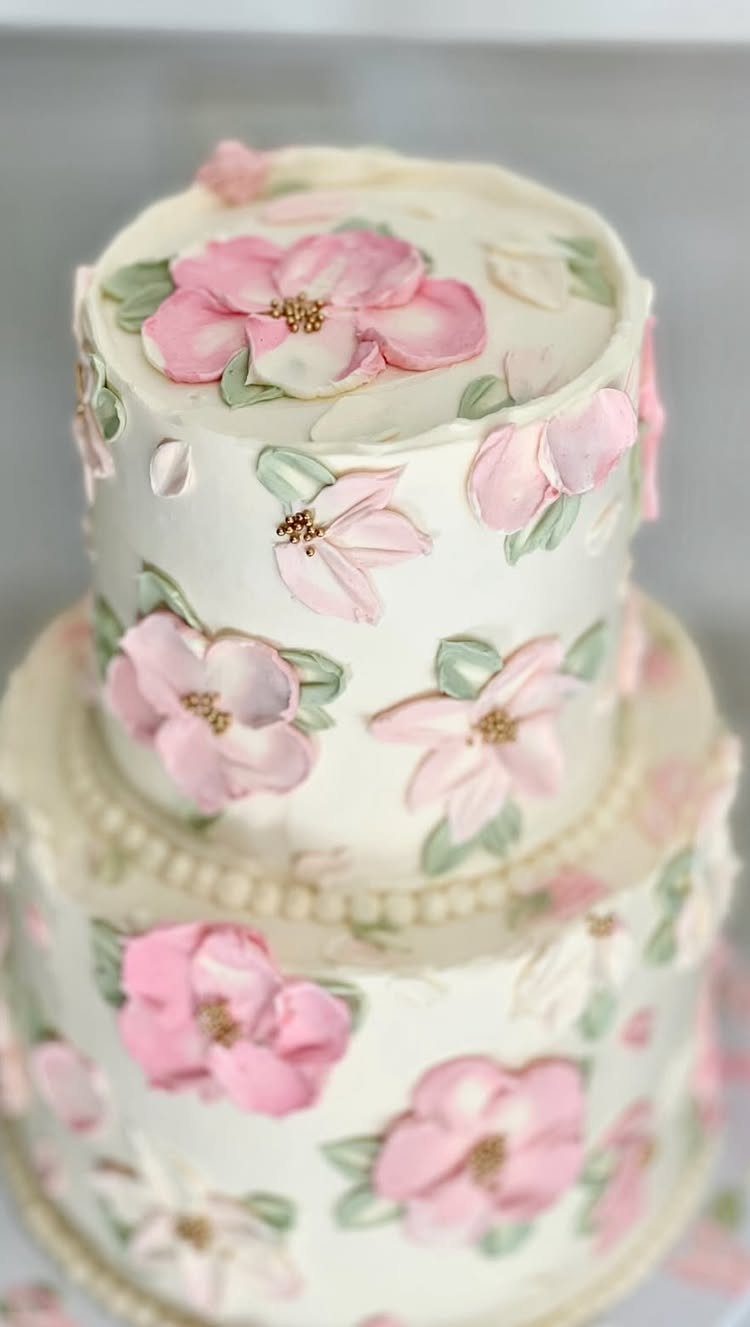 Two-tiered white buttercream cake decorated with large pink floral patterns and green leaves.
