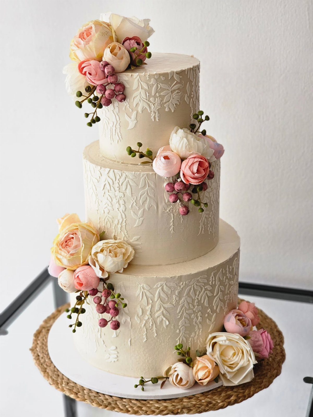Three-tier white buttercream wedding cake decorated with clusters of pink and peach roses and small berries.