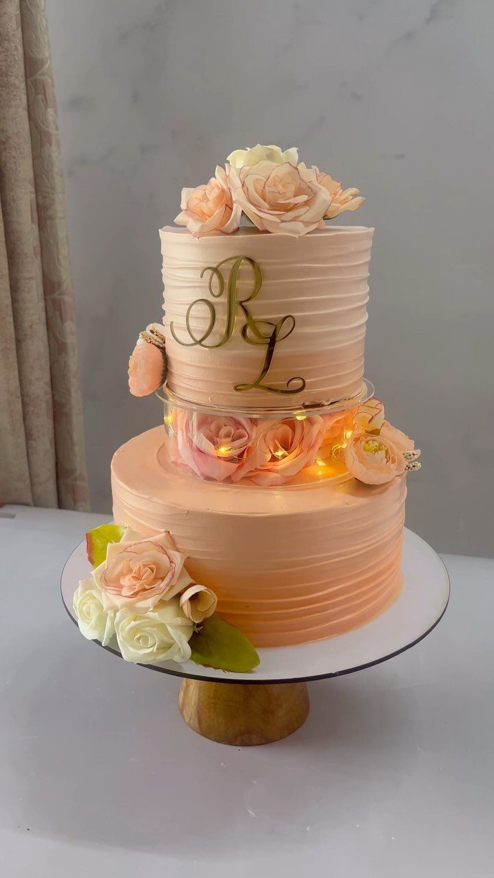 Two-tier peach buttercream wedding cake decorated with roses and fairy lights between the tiers.